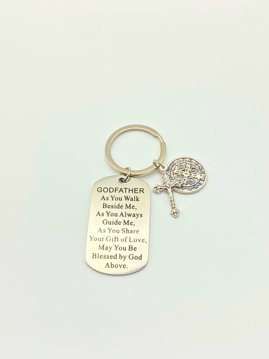 A silver-toned metal keychain featuring a dog tag engraved with a Godfather quote, a small cross, and a round St. Benedict charm, displayed on a clean off-white background.